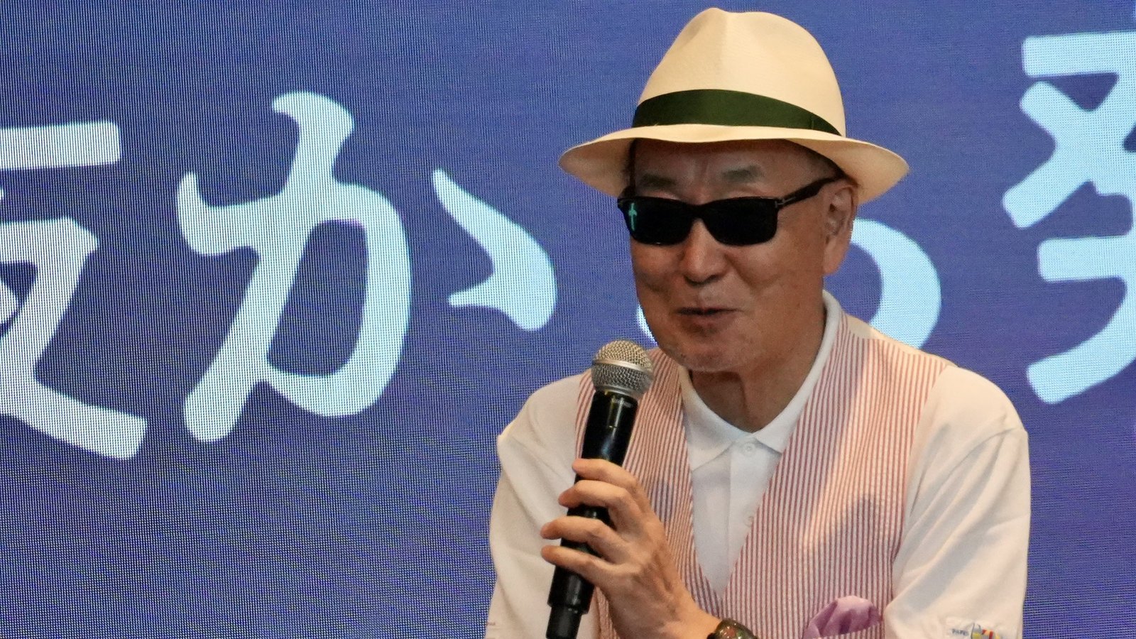 Oji Hiroi speaks on stage while holding a microphone, wearing a hat and sunglasses
