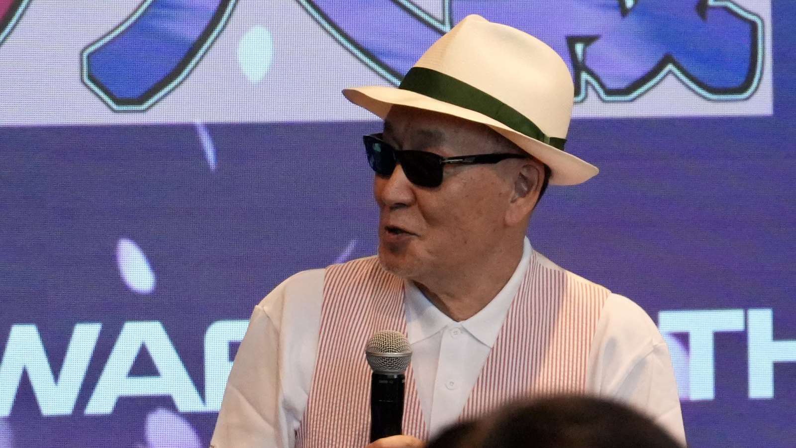 Oji Hiroi speaks in profile in front of a purple 30th-anniversary visual