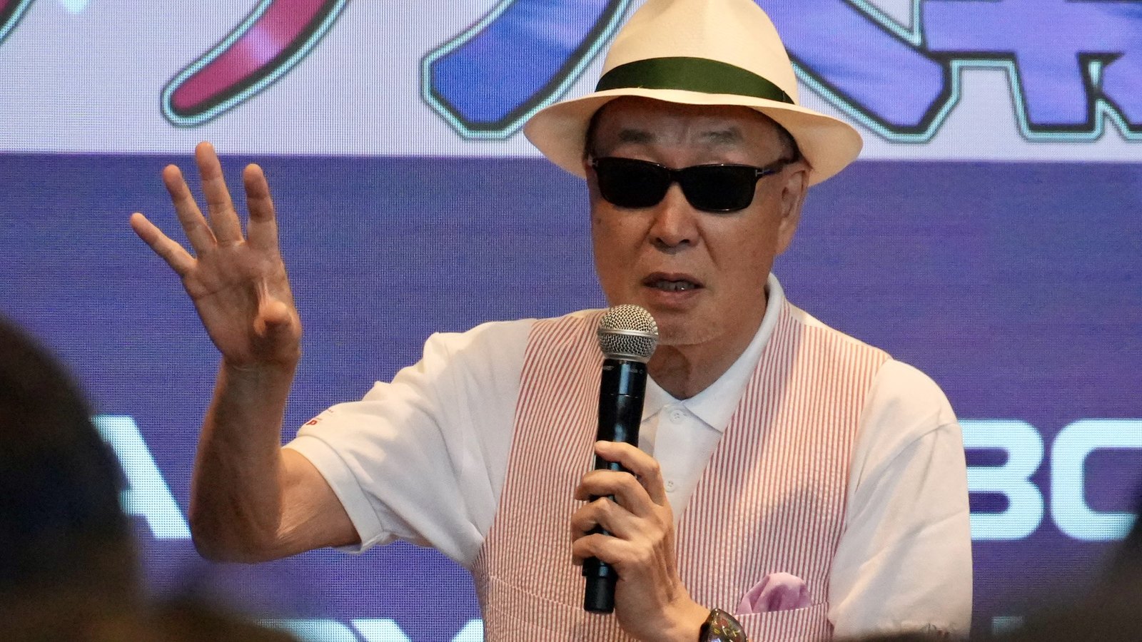 Oji Hiroi speaks while holding a microphone, one hand raised and open